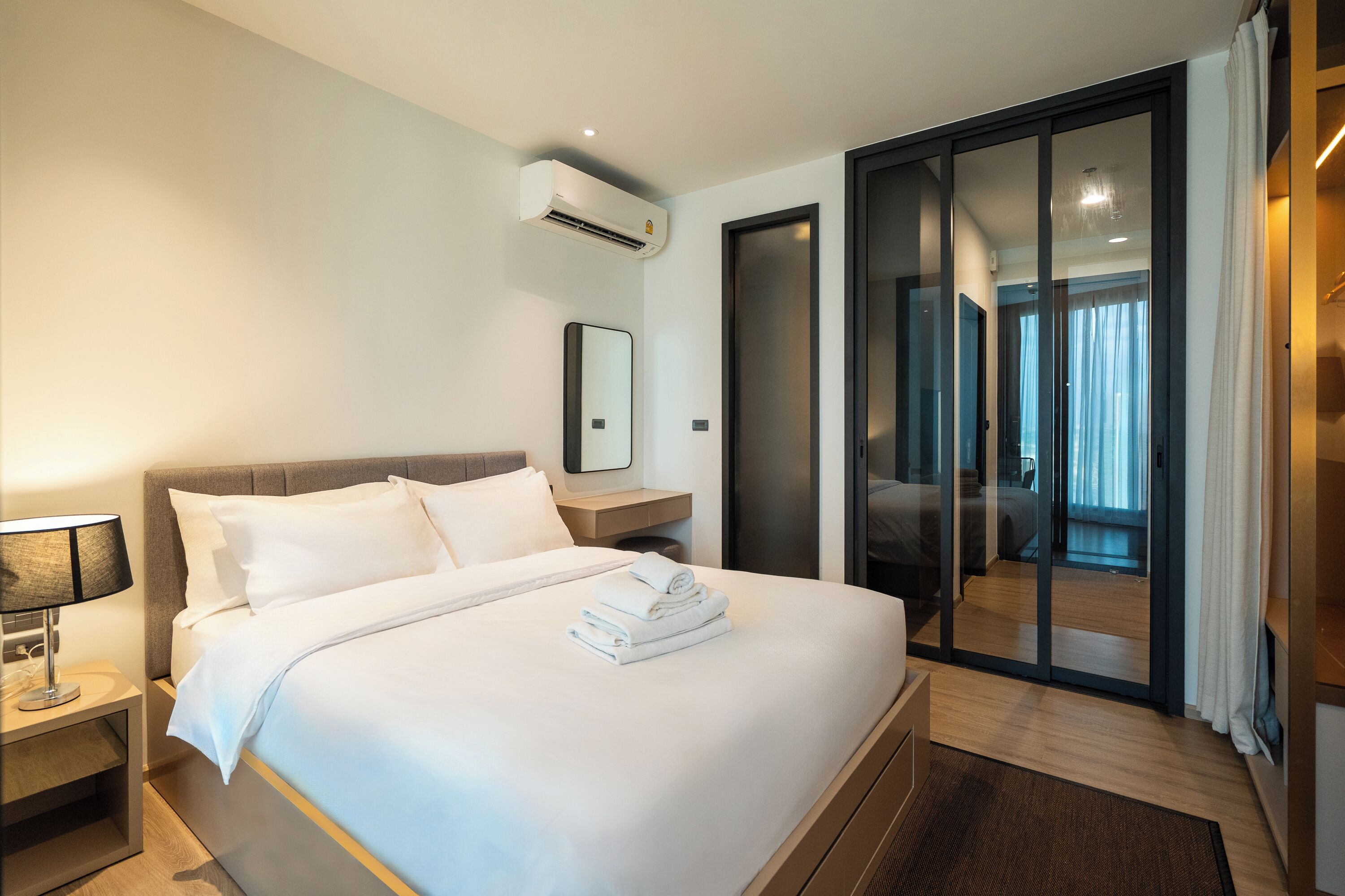 Ein modernes Hotelzimmer mit einem großen Bett, einem Nachttisch mit Lampe, einem Spiegel und einer Schiebeglastür, die auf einen Balkon führt.