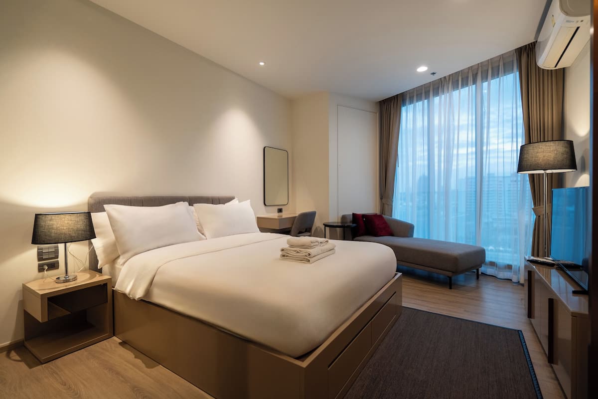 A modern hotel room with a large bed, a sofa, a TV, and a city view.