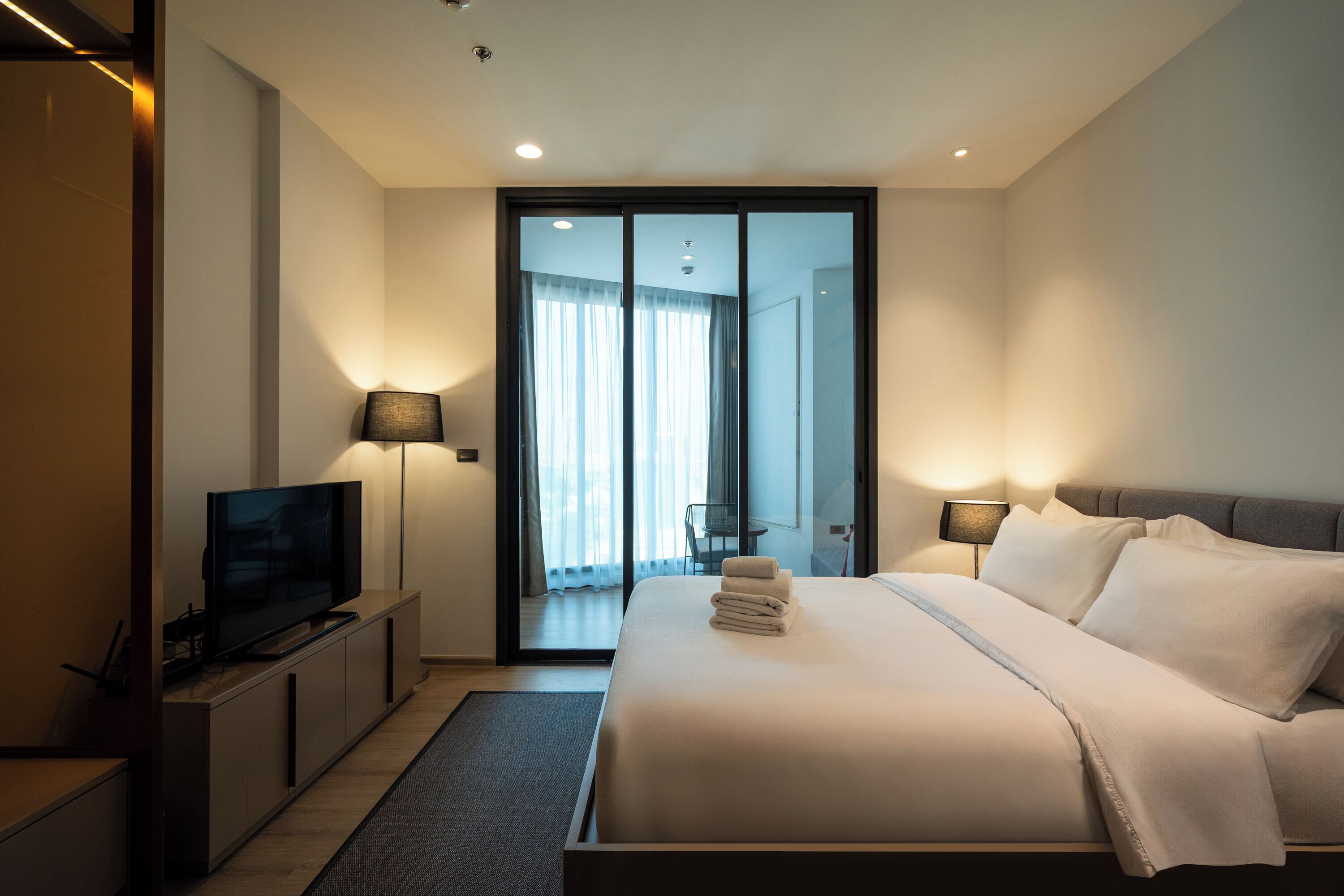 A modern hotel room with a large bed, a flat-screen TV, a desk, and a sliding door leading to a balcony.