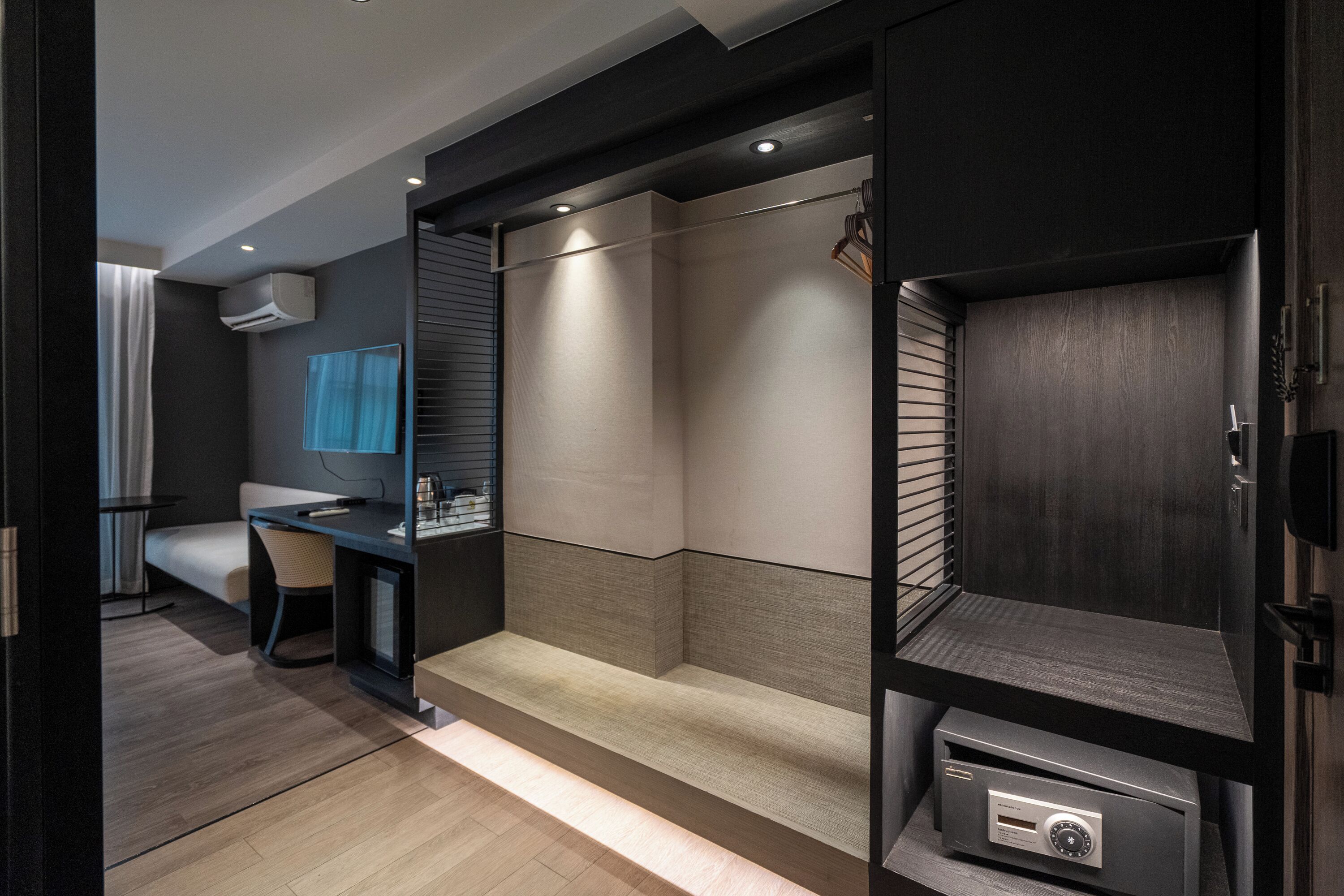 A modern hotel room with a dark wood finish, a small desk with a chair, a large mirror, and a built-in wardrobe with a mirror.