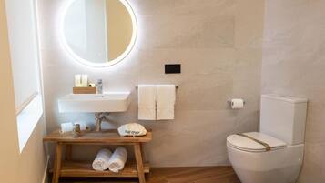 A modern bathroom with a round mirror, white sink, and toilet. There is a wooden shelf with towels and a hair dryer.