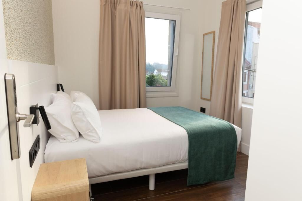 Un dormitorio con cama, mesita de noche, ventana con cortinas y vistas a edificios.
