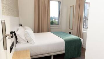 Un dormitorio con cama, mesita de noche, ventana con cortinas y vistas a edificios.