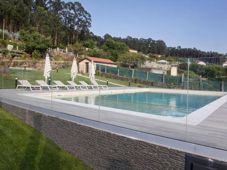 Una piscina rectangular con agua azul cristalina, rodeada por una terraza de madera y un muro de piedra. Hay sillas de descanso y sombrillas a lo largo de la piscina.
