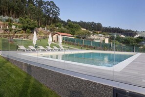 A rectangular swimming pool with clear blue water, surrounded by a wooden deck and a stone wall. There are lounge chairs and umbrellas along the poolside.