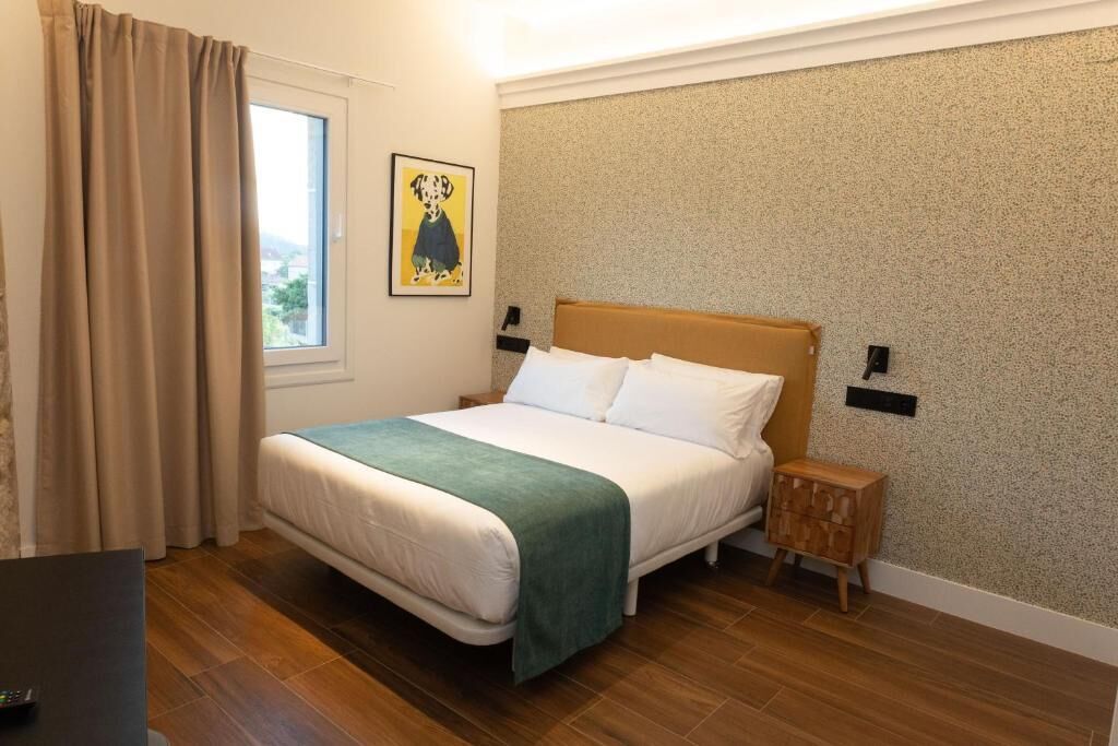 A neatly arranged hotel room with a bed, a small bedside table, a window with curtains, and a wall-mounted light fixture.
