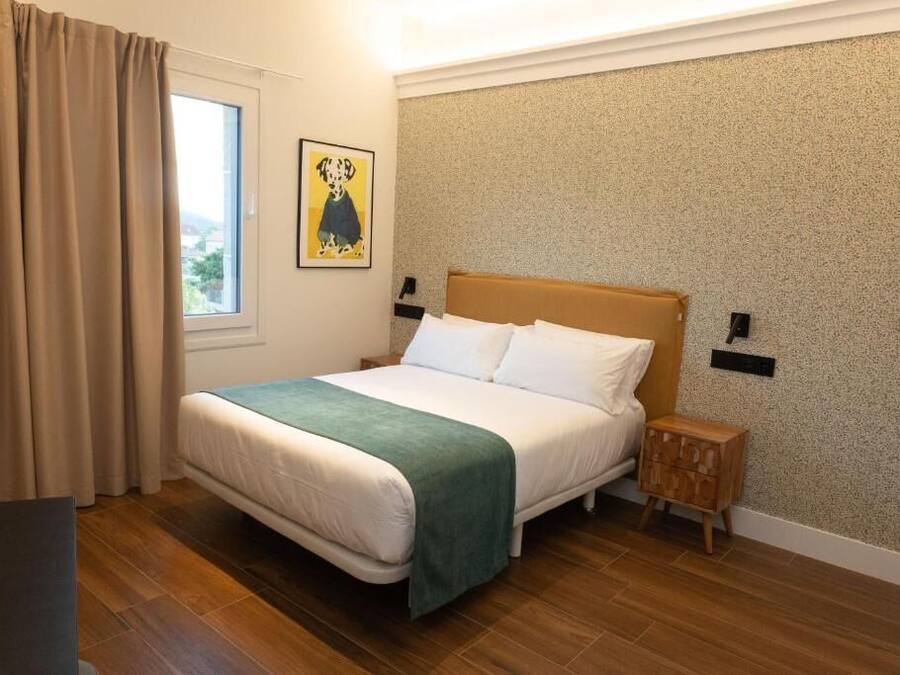 Habitación de hotel ordenada con cama, mesita de noche, ventana con cortinas e iluminación fijada a la pared.