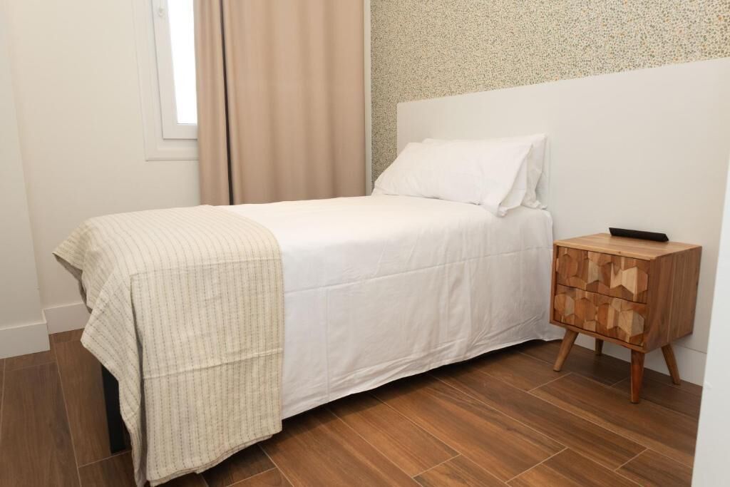 Un dormitorio con una cama, una mesita de noche de madera, una ventana con cortinas y un muro con textura.