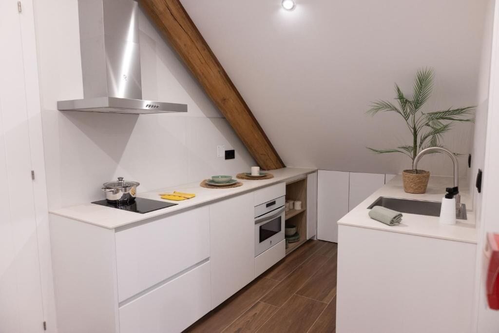 A modern kitchen with white cabinetry, a stainless steel range hood, a wooden beam ceiling, and a small potted plant.