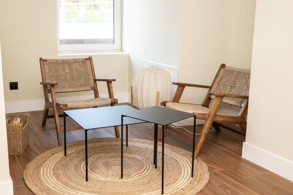A room with two wooden chairs and a small table, a woven rug, and a window with a view of greenery.