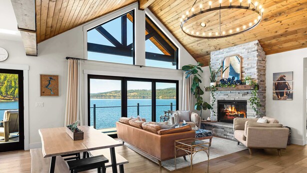 Amplio salón con un ventanal grande con vistas a un lago, chimenea y techo de madera alto.