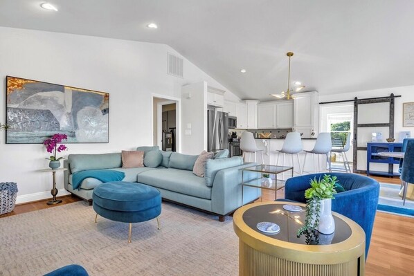 A modern living room with a sofa, ottoman, and coffee table, a dining area with chairs, and a kitchen with white cabinets and appliances.