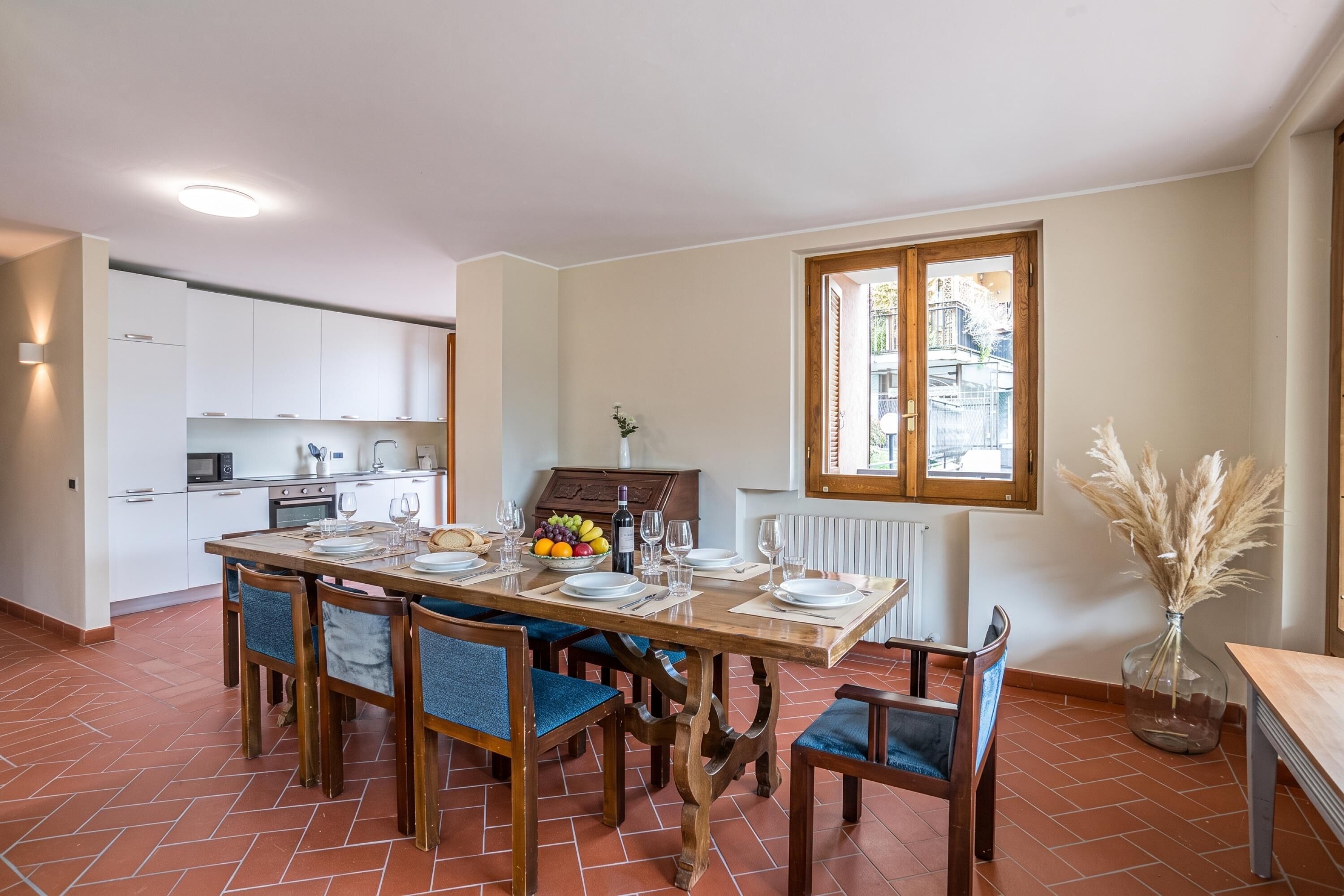 A dining area with a wooden table set for six, surrounded by blue upholstered chairs. A kitchen with white cabinets and a window is visible in the background.