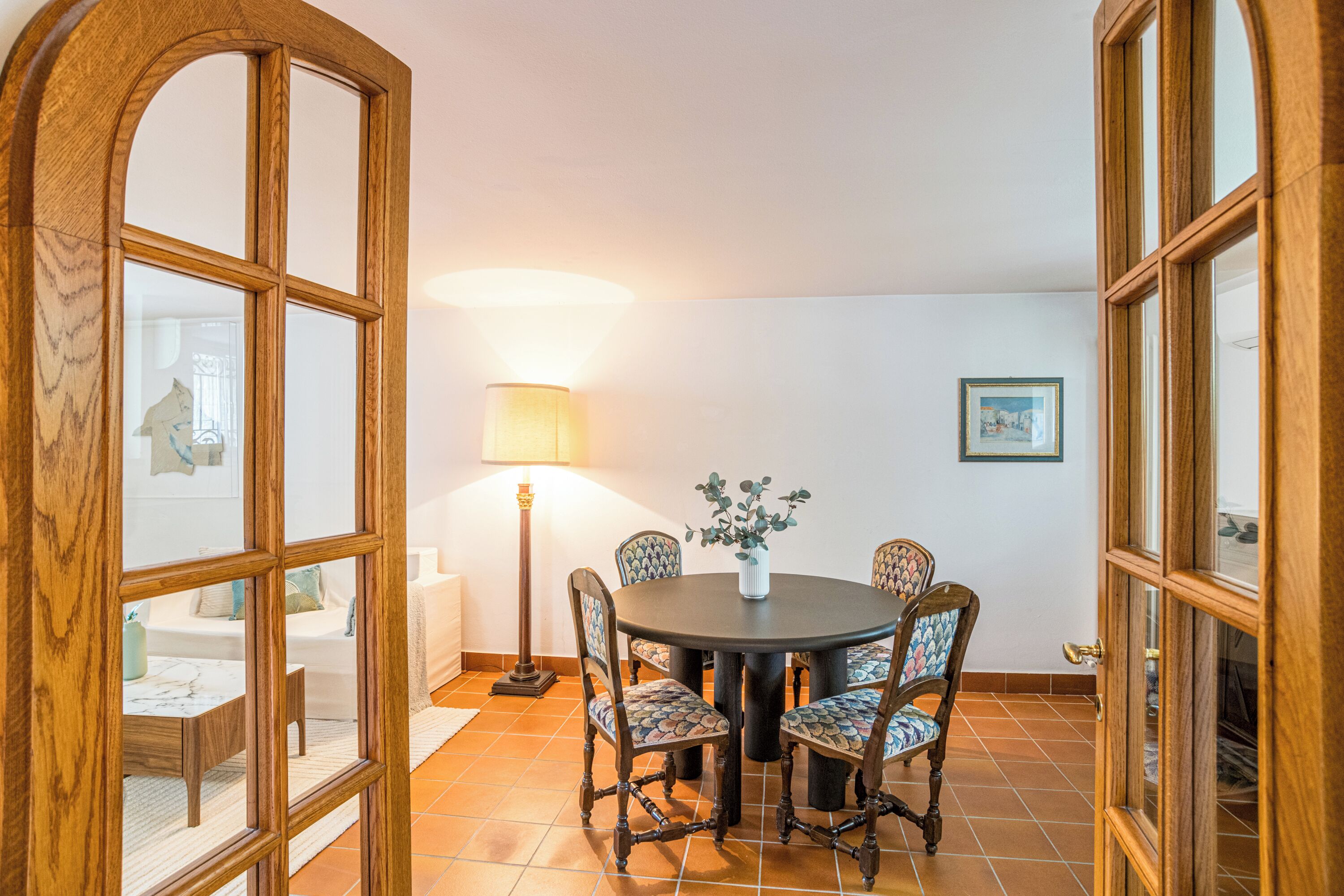 A dining area with a round table, chairs, a lamp, and a vase of flowers.