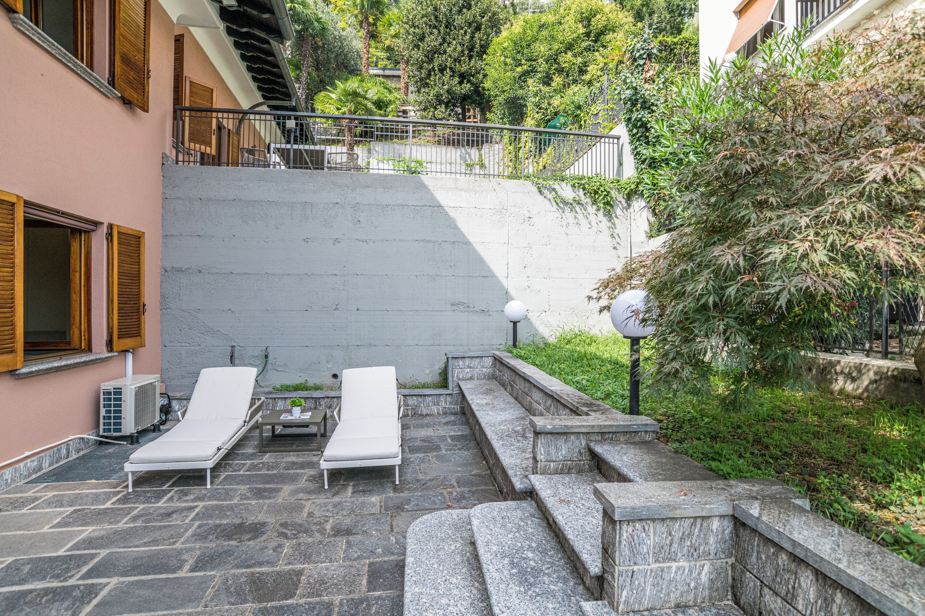A stone patio with two white lounge chairs and a small table, surrounded by greenery and a concrete wall.