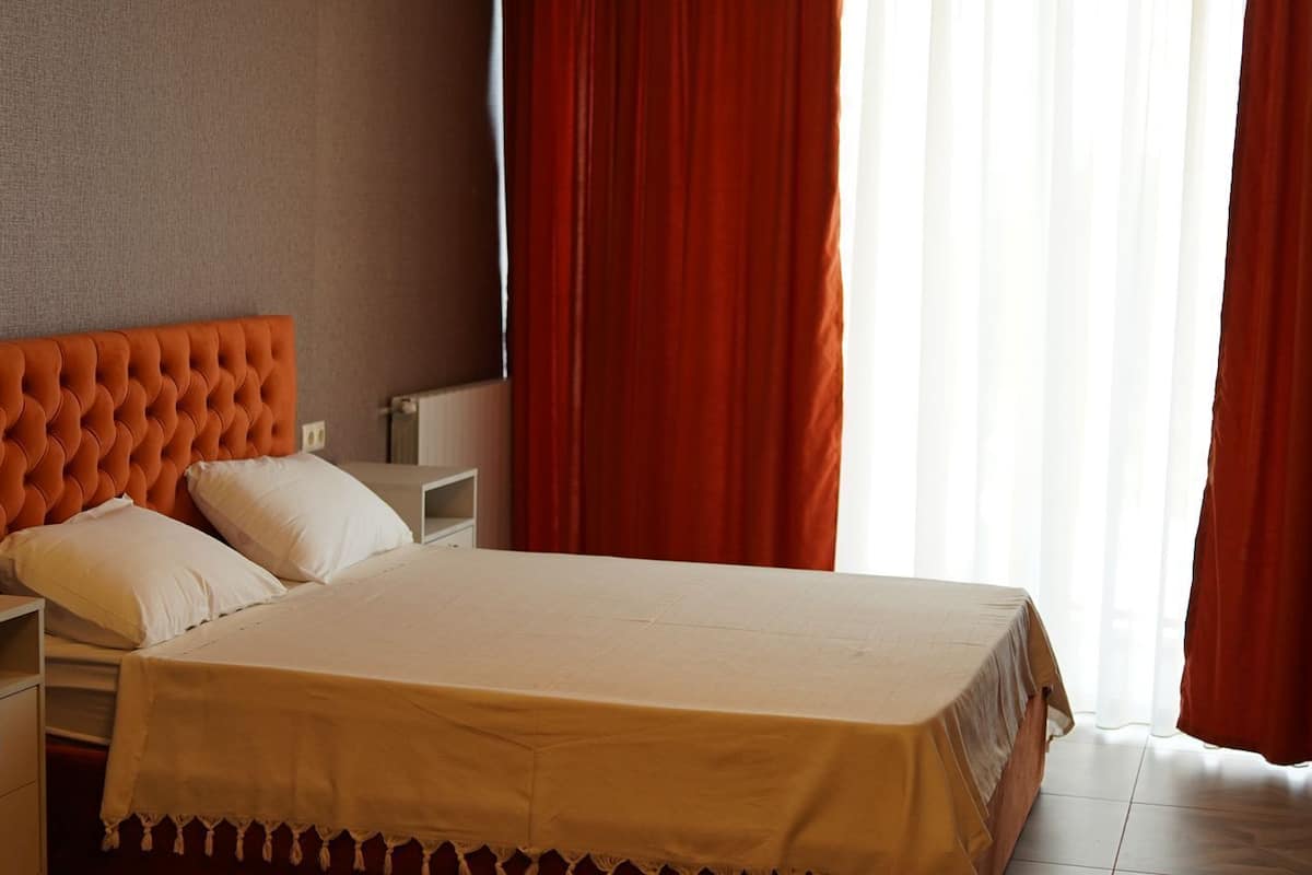 A hotel room with a large bed, a nightstand, a dresser, and a window with red curtains.
