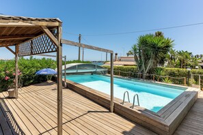 A wooden deck with a swimming pool, surrounded by greenery and a clear sky.
