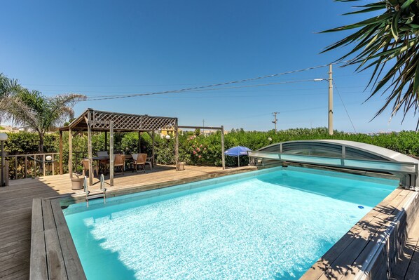 A swimming pool with a wooden deck, a covered seating area, and a glass bridge.