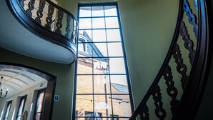 Un grand escalier orné d’un lustre, une grande fenêtre donnant sur un immeuble et une porte avec un numéro.