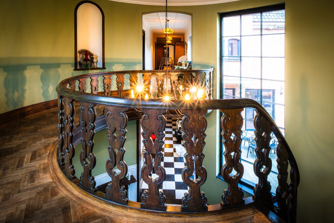 A grand staircase with ornate railings and a checkered floor, leading to a bright room with a chandelier.