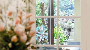 A dining area with a table set for two, a vase of flowers, and a view of an outdoor garden through a glass door.