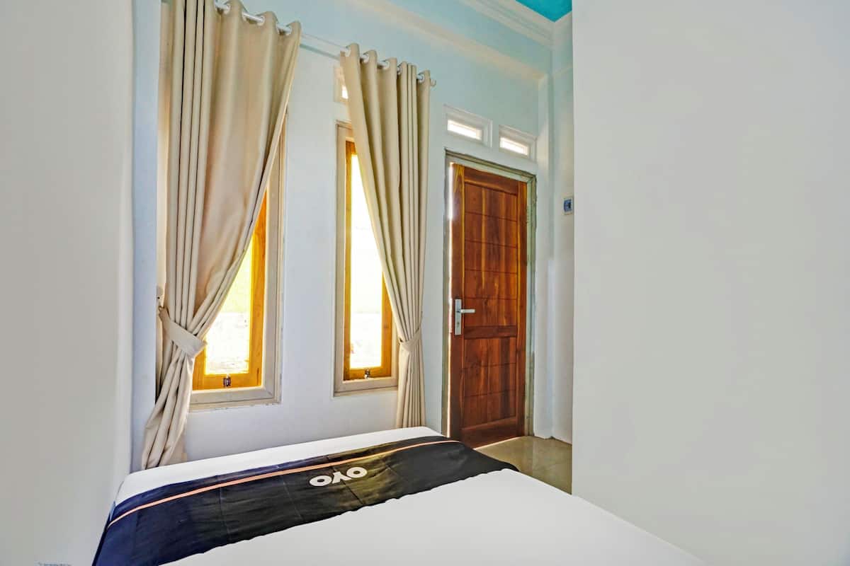 A bedroom with a bed, two windows with curtains, a wooden door, and a light blue wall.