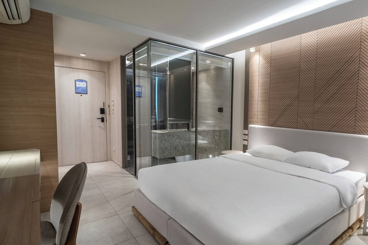 A modern hotel room with a large bed, a glass-enclosed shower, and a wooden panel wall.