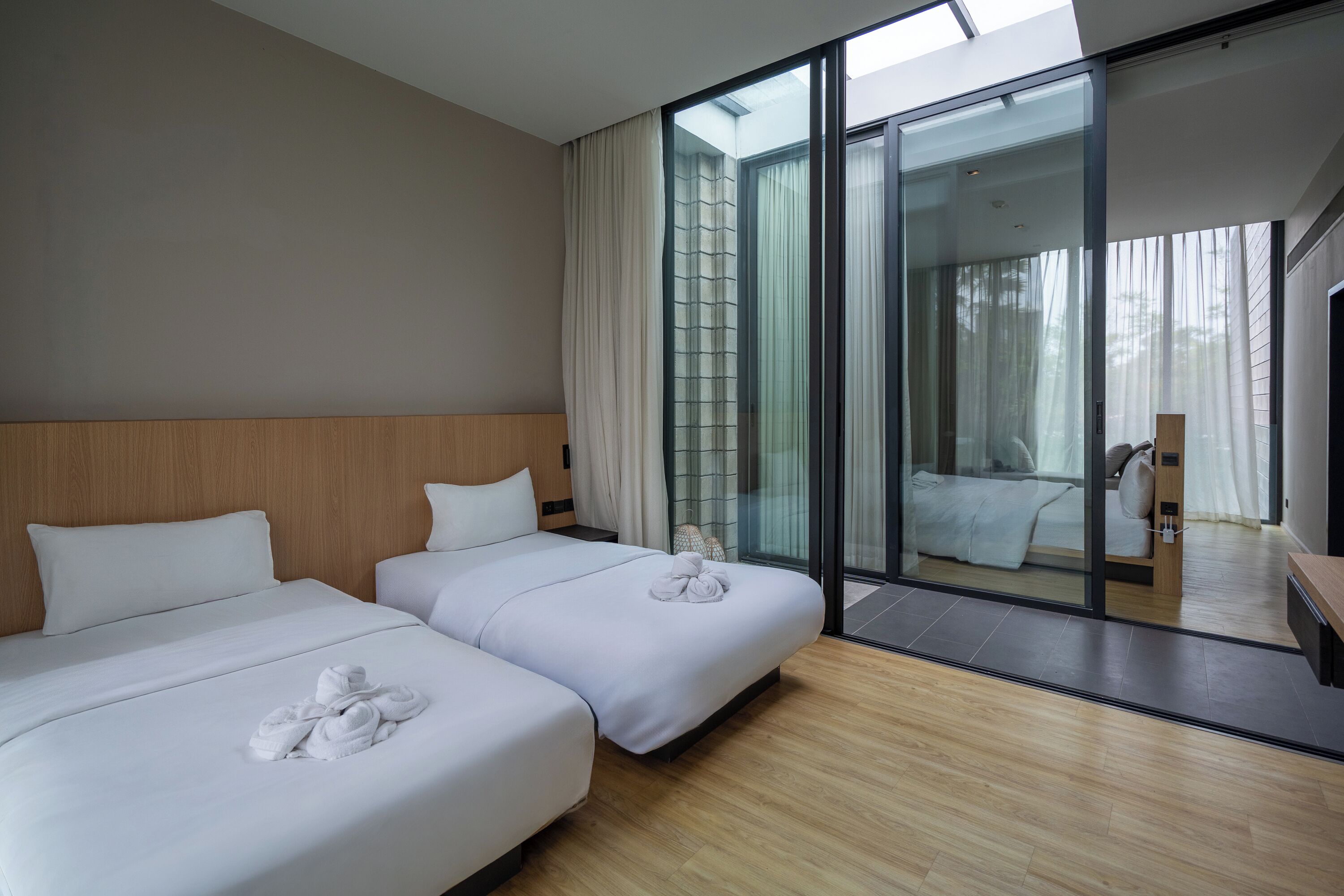 A hotel room with two beds, a wooden headboard, a large sliding glass door leading to a balcony, and a wooden floor.