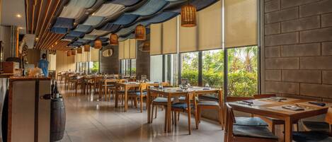 Um restaurante espaçoso com mesas e cadeiras de madeira, grandes janelas com vista para o verde e um design de teto moderno com lanternas pendentes.