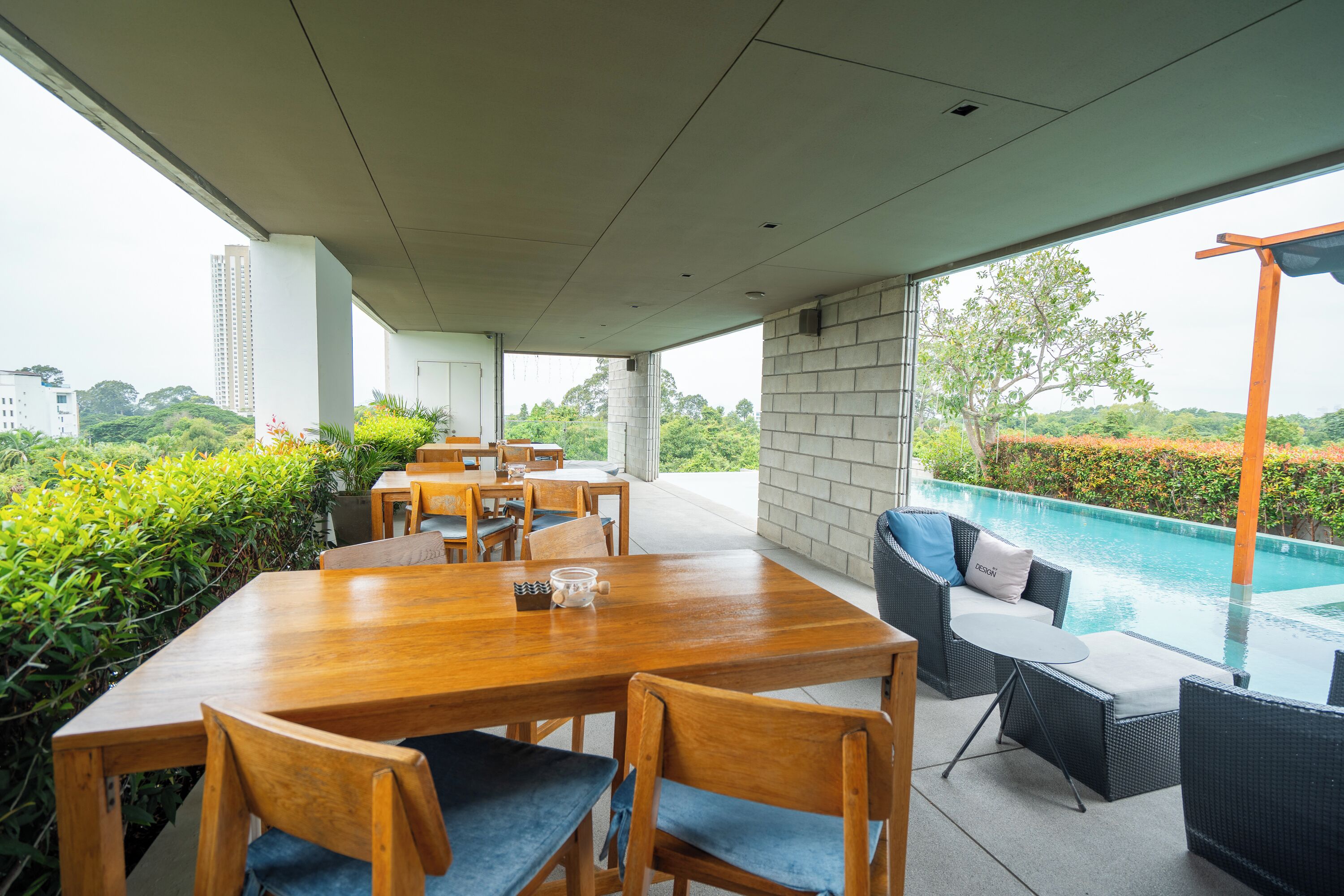 A covered outdoor dining area with wooden tables and chairs, a pool in the background, and a view of greenery and buildings.
