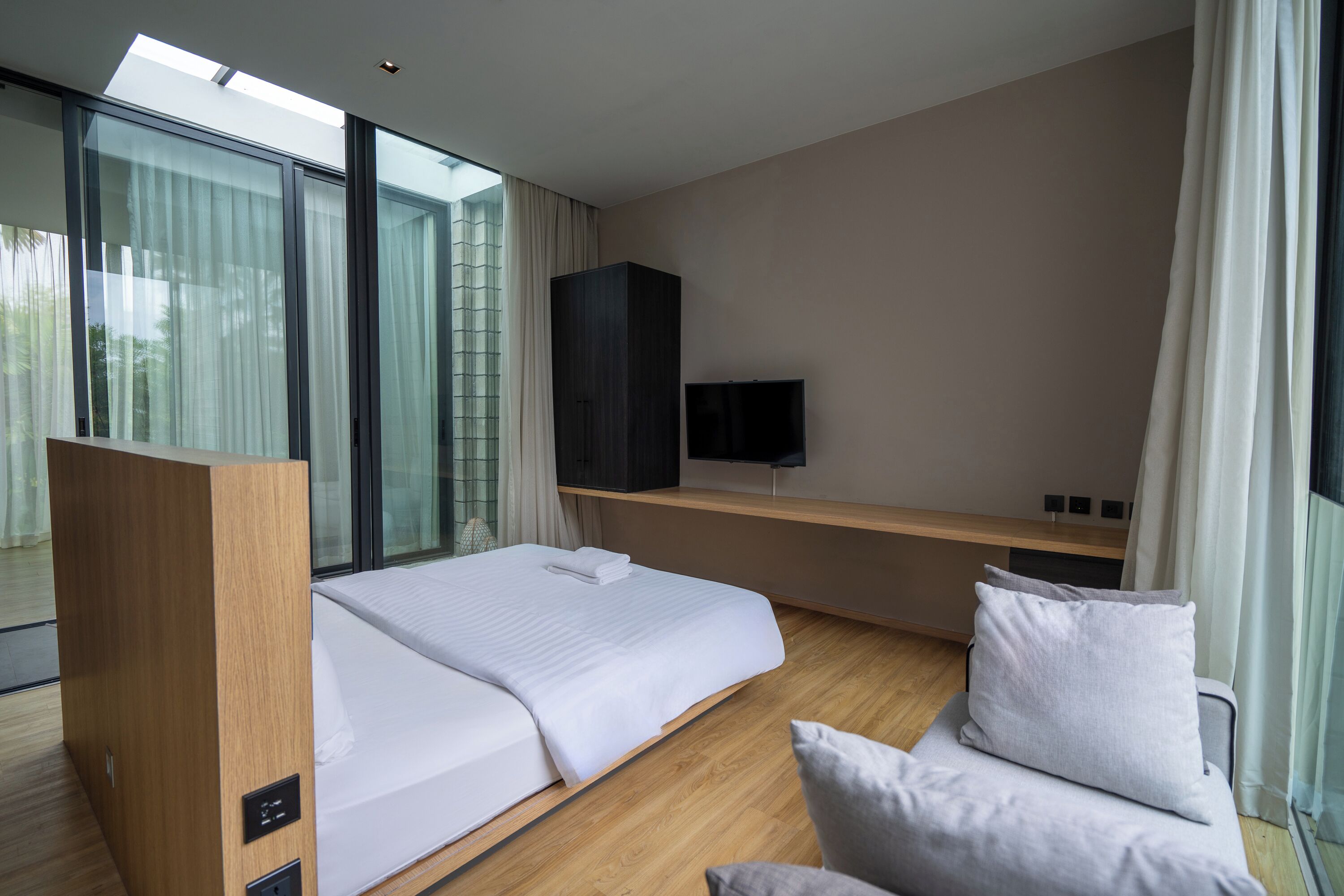 Una habitación de hotel moderna con una cama grande, un armario de madera, un televisor sobre una estantería de madera y un amplio ventanal con cortinas.