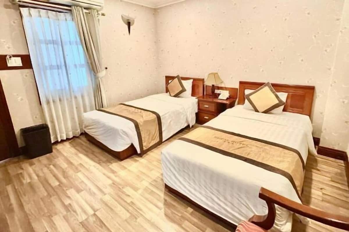 Habitación de hotel con dos camas, piso de madera, mesita de noche, lámpara y ventana con cortinas.
