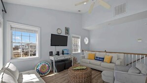 A spacious living room with a white sofa, a grey armchair, a wicker ottoman, and a flat-screen TV mounted on the wall. - Oceanfront Retreat, Private Beach, Stunning Views (North Topsail Beach)
