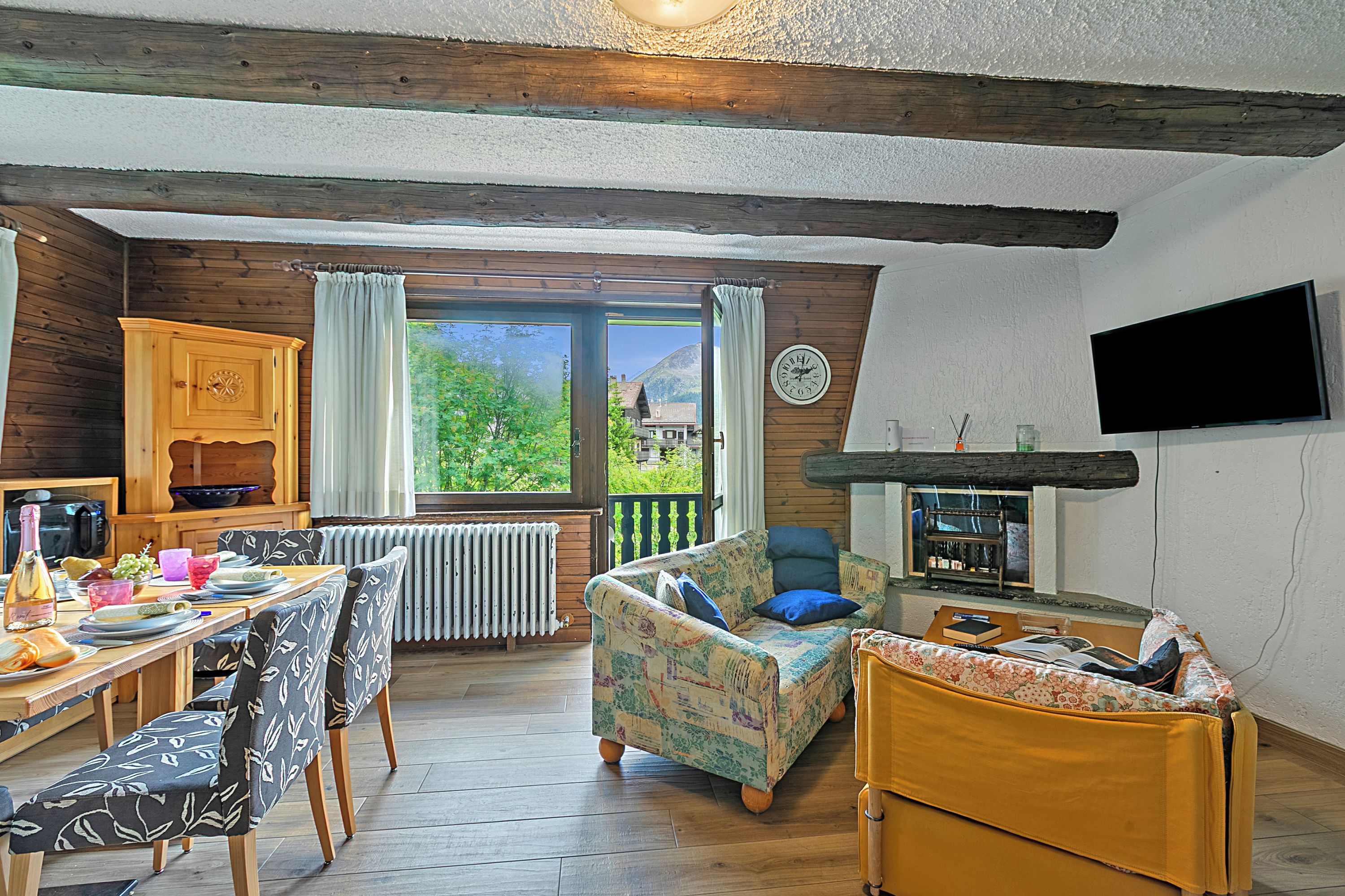 A cozy room with a wooden dining table, chairs, a sofa, a television, and a view of greenery outside.