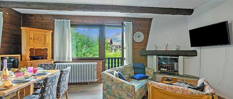A cozy room with a wooden dining table, chairs, a sofa, a television, and a view of greenery outside.