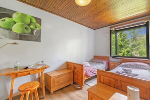 Un dormitorio con cama de madera, un escritorio con taburete, una mesita de noche y una ventana con vistas a la vegetación.