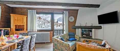 A cozy living room with a wooden ceiling, a dining area with a table and chairs, a sofa, and a television mounted on the wall.