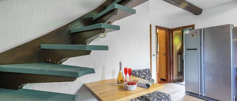 Una escalera con peldaños verdes, una mesa de madera con un cuenco de fruta y dos vasos rojos, una silla estampada y un refrigerador en una habitación con vigas de madera y una pared blanca.