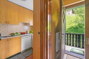 Eine Küche mit Holzausstattung, einem weißen Geschirrspüler und einem Fenster mit Blick auf einen Balkon, der viel Grün und ein Gebäude im Hintergrund zeigt.