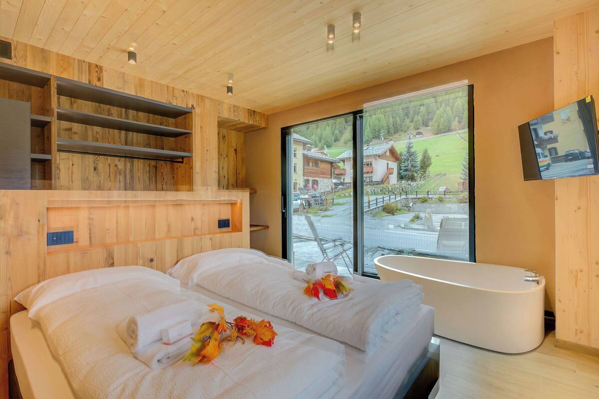 A modern hotel room with a wooden ceiling, a large window with a view of the outside, a bathtub, and a flat-screen TV mounted on the wall.