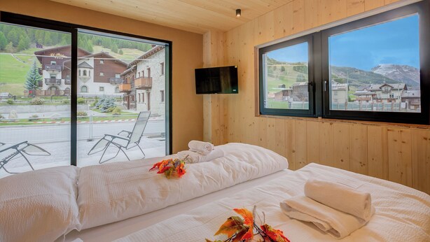 Ein Hotelzimmer mit einem großen Bett, einem an der Wand montierten Fernseher und Blick auf ein Bergdorf durch große Fenster.