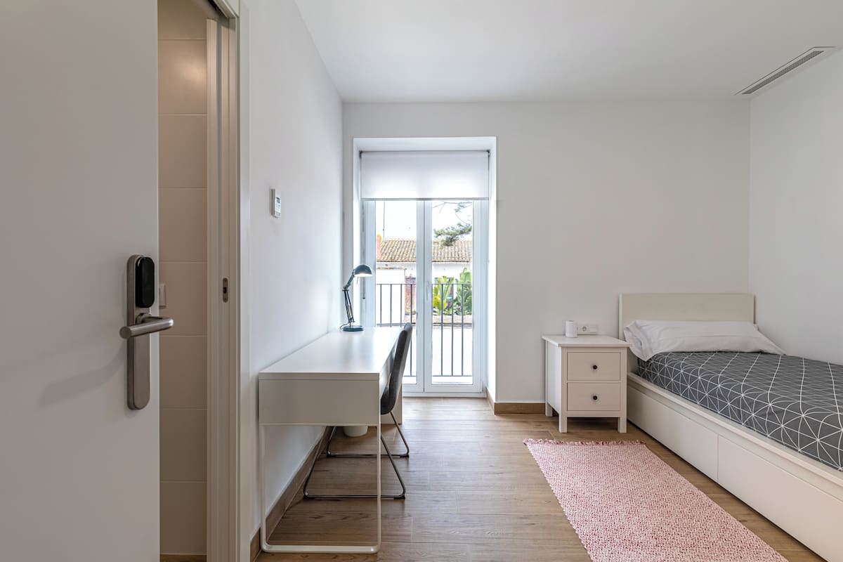 A bedroom with a bed, bedside table, desk, chair, and a view through a sliding door to an outdoor area.