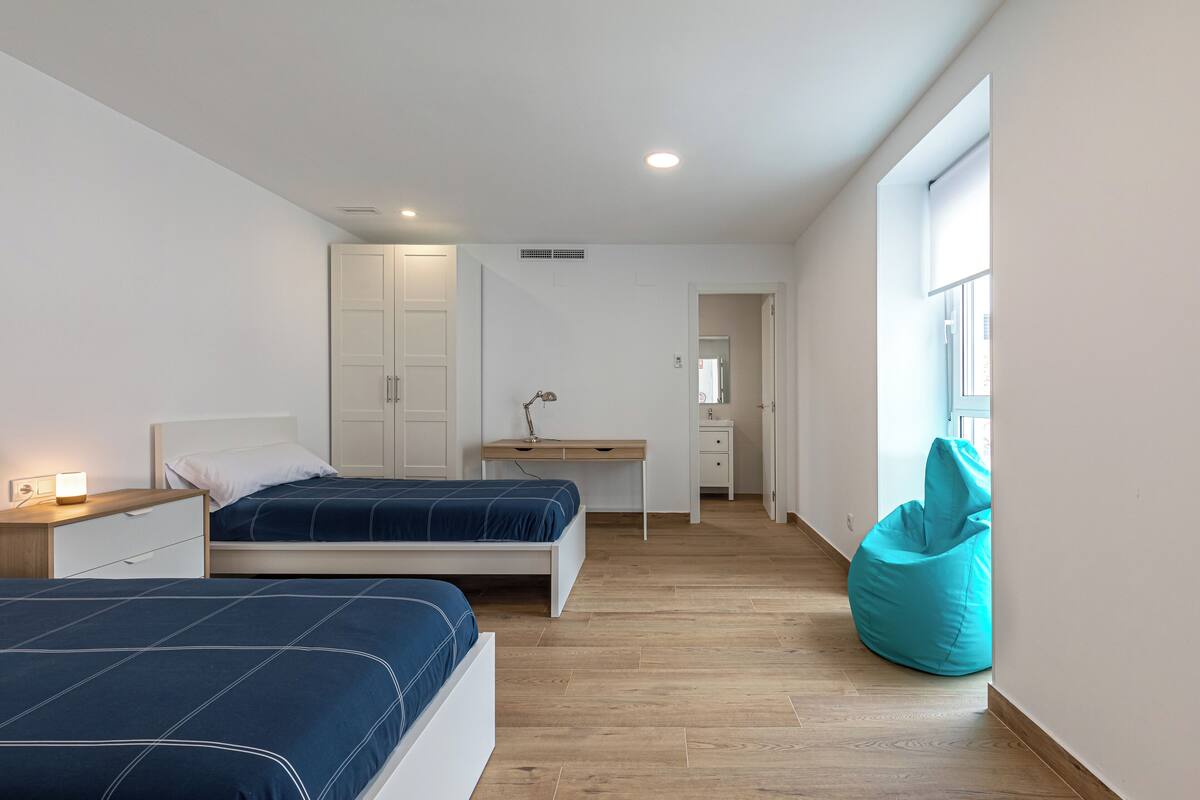 A bedroom with two beds, a desk, a chair, a bean bag, and a window.