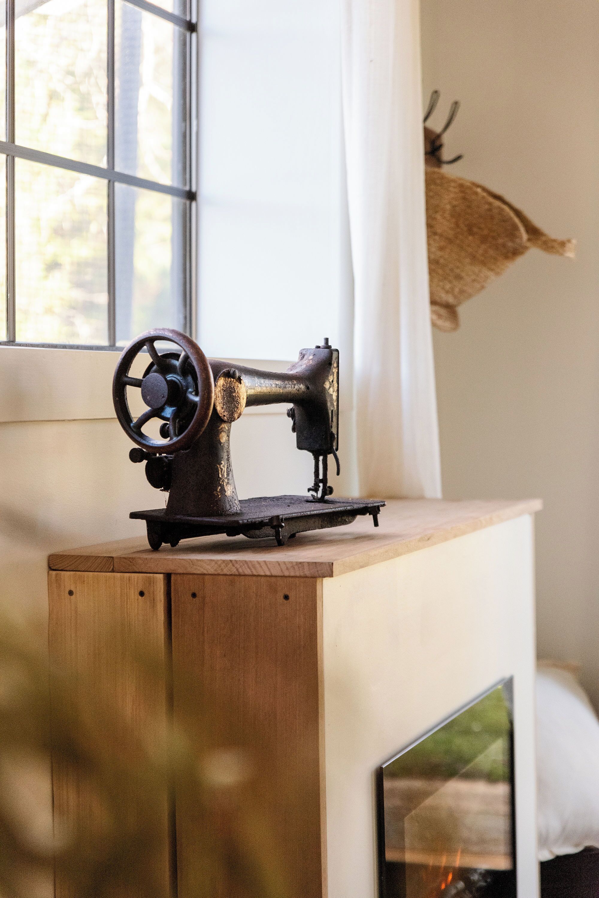 A vintage sewing machine on a wooden table near a window with a deer head mounted on the wall.