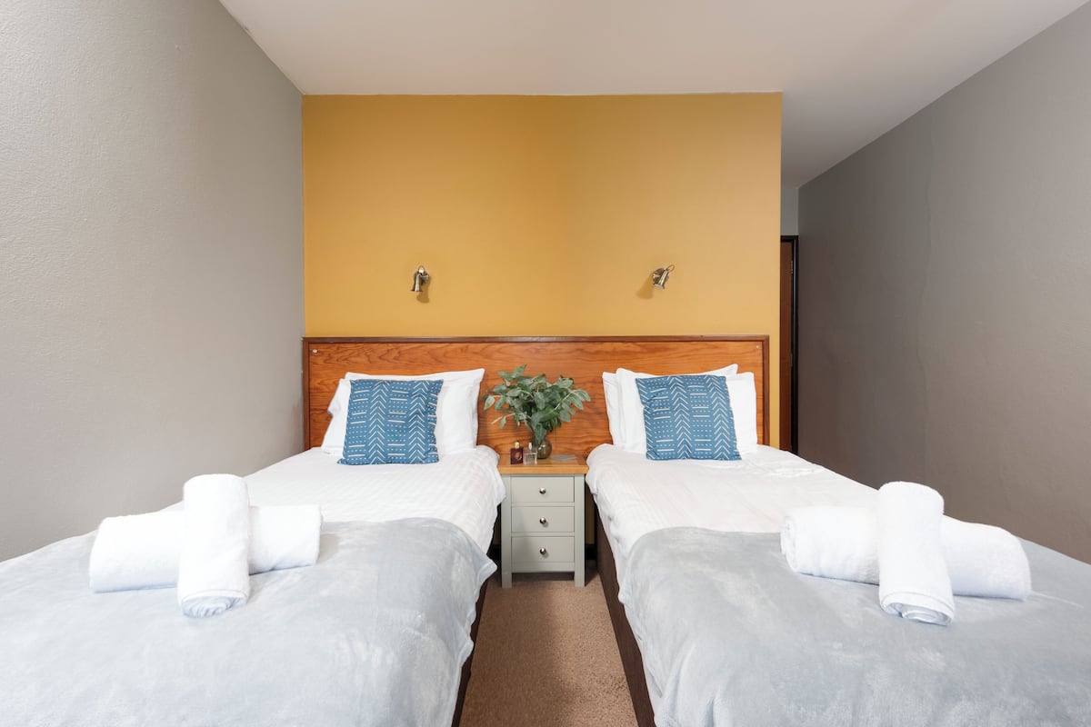 A hotel room with two beds, a wooden headboard, a small bedside table with a plant, and a beige wall.