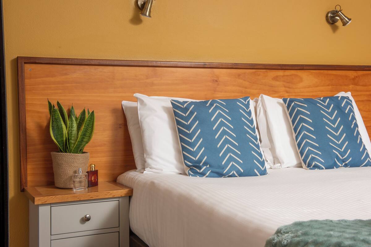A neatly made bed with white and blue patterned pillows, a small potted plant, and a bedside table with a bottle and a container.