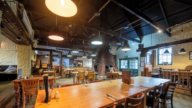 Un restaurante espacioso con techos altos, vigas a la vista y un horno de ladrillo grande. Las mesas están preparadas con cubiertos, y el espacio está bien iluminado con lámparas colgantes.