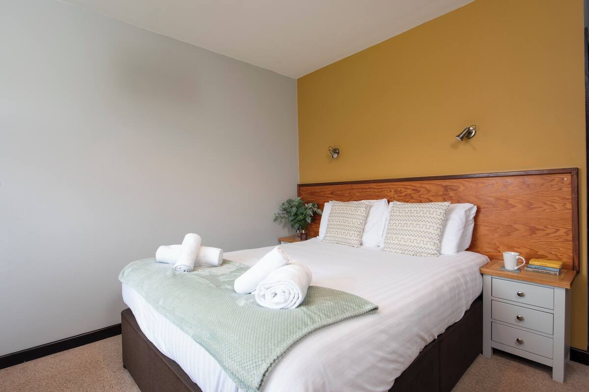 A neatly made bed with rolled towels, a bedside table with a cup and book, and a nightstand with a plant.