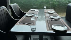 Mesa de jantar posta para dois, cadeiras e vista para o verde lá fora, com uma janela embaçada.