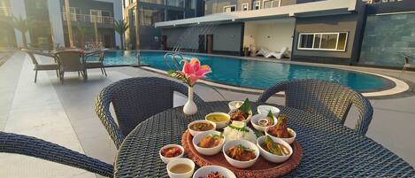 A dining table with various dishes, a pool, and modern buildings in the background.